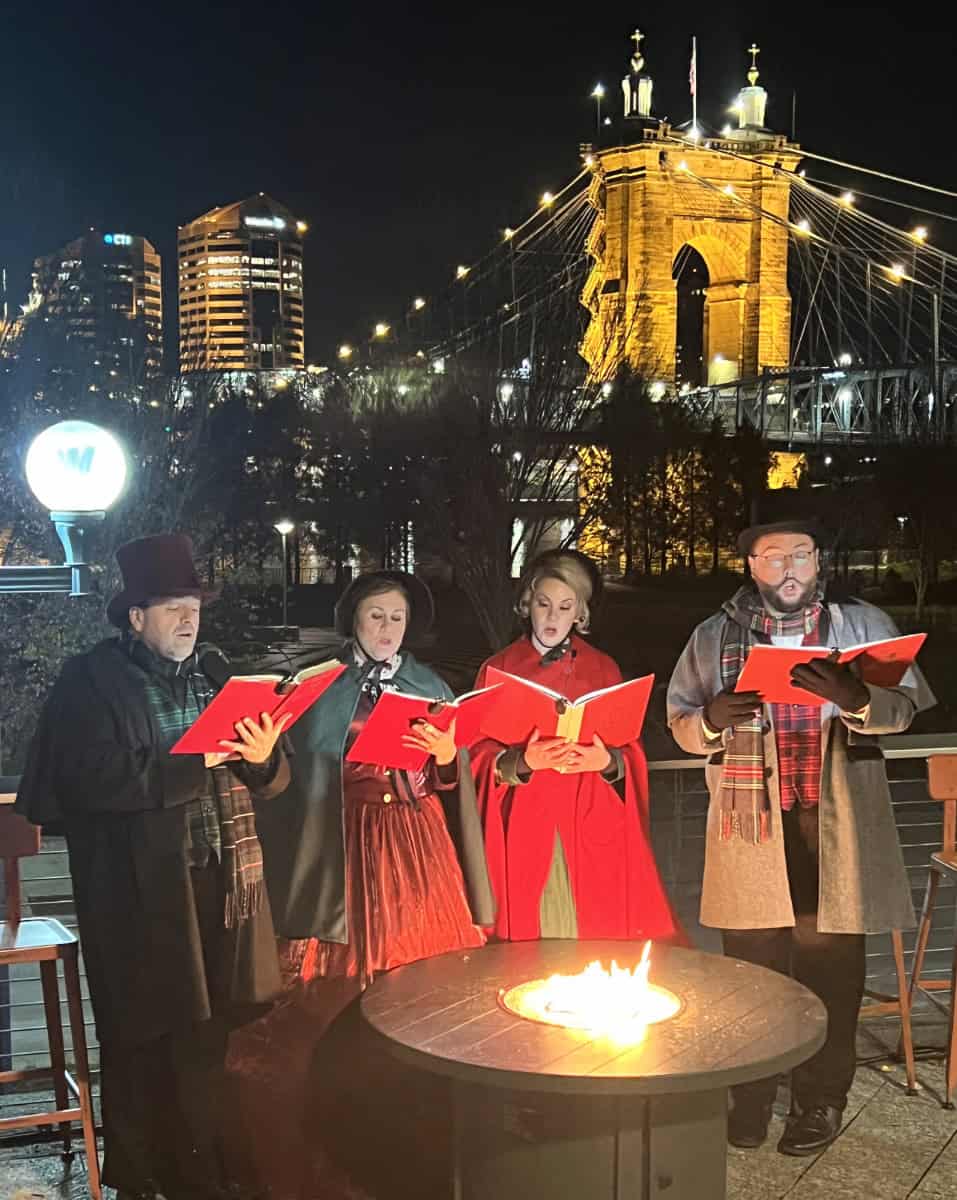 carolers signing along the Ohio River with the Roebling Bridge in the background