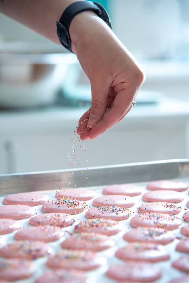 macarons on a baking sheet with a hand sprinkling sprinkles over them